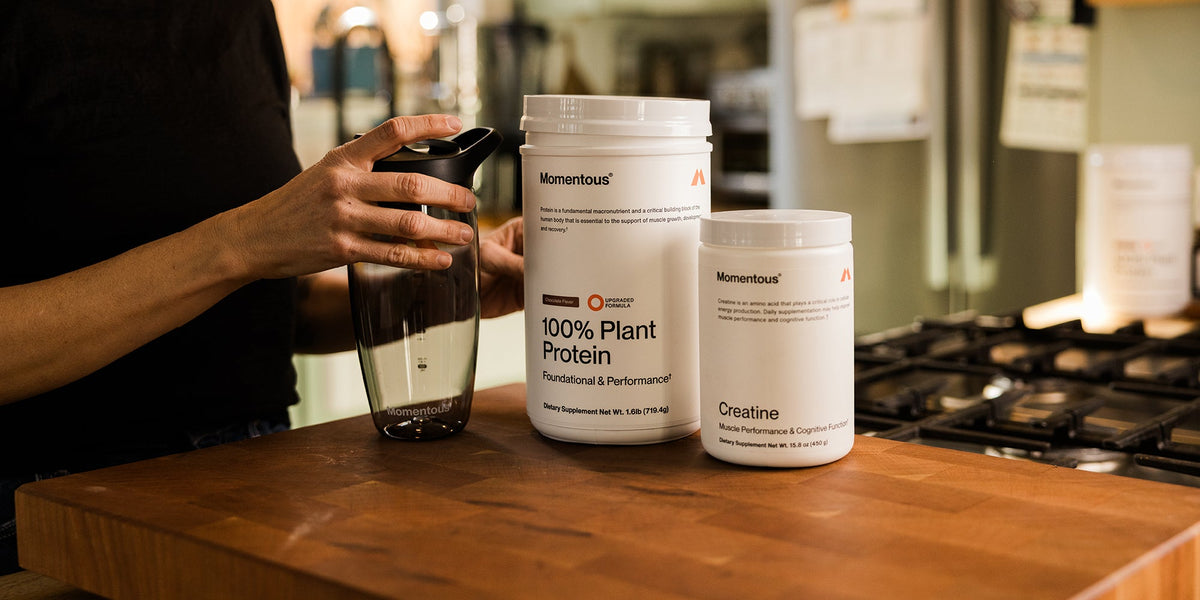 a tub of Momentous Creatine and a tub of Momentous Plant Protein next to a shaker and a person's hand