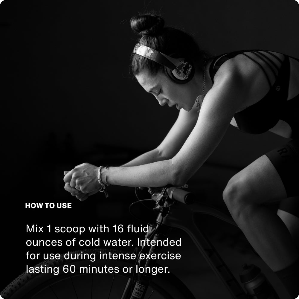 Black-and-white photo of a woman wearing Red Bull-branded headphones, leaning forward while riding an indoor bike trainer. She appears focused and exerting effort. White text on the left side reads: 'HOW TO USE — Mix 1 scoop with 16 fluid ounces of cold water. Intended for use during intense exercise lasting 60 minutes or longer.'