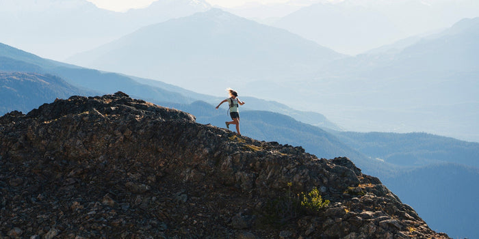 A female runner runs down a mountain trail with mountains behind her