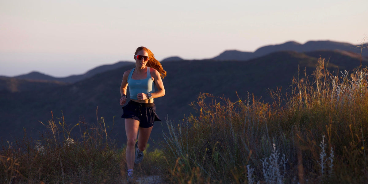 A young woman runs on a grassy trail with mountains behind her