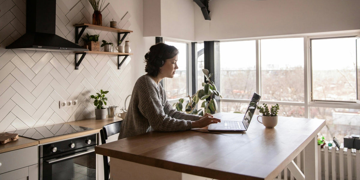 A woman sits at her kitchen island working on her laptop with headphones on
