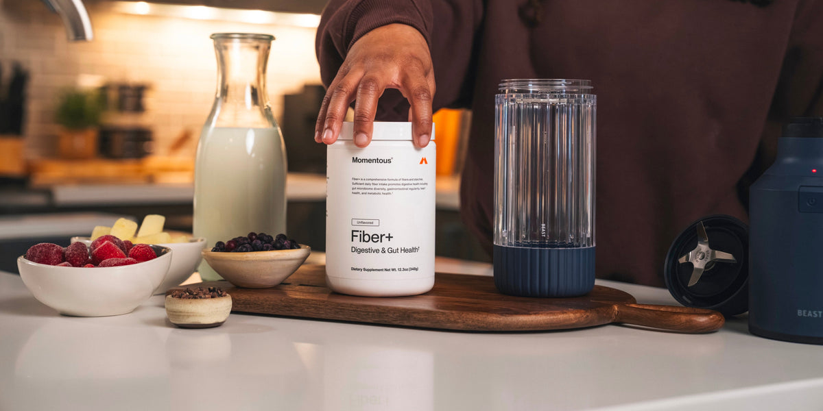 A tub of Momentous Fiber+ surrounded by fruit and milk and a blender