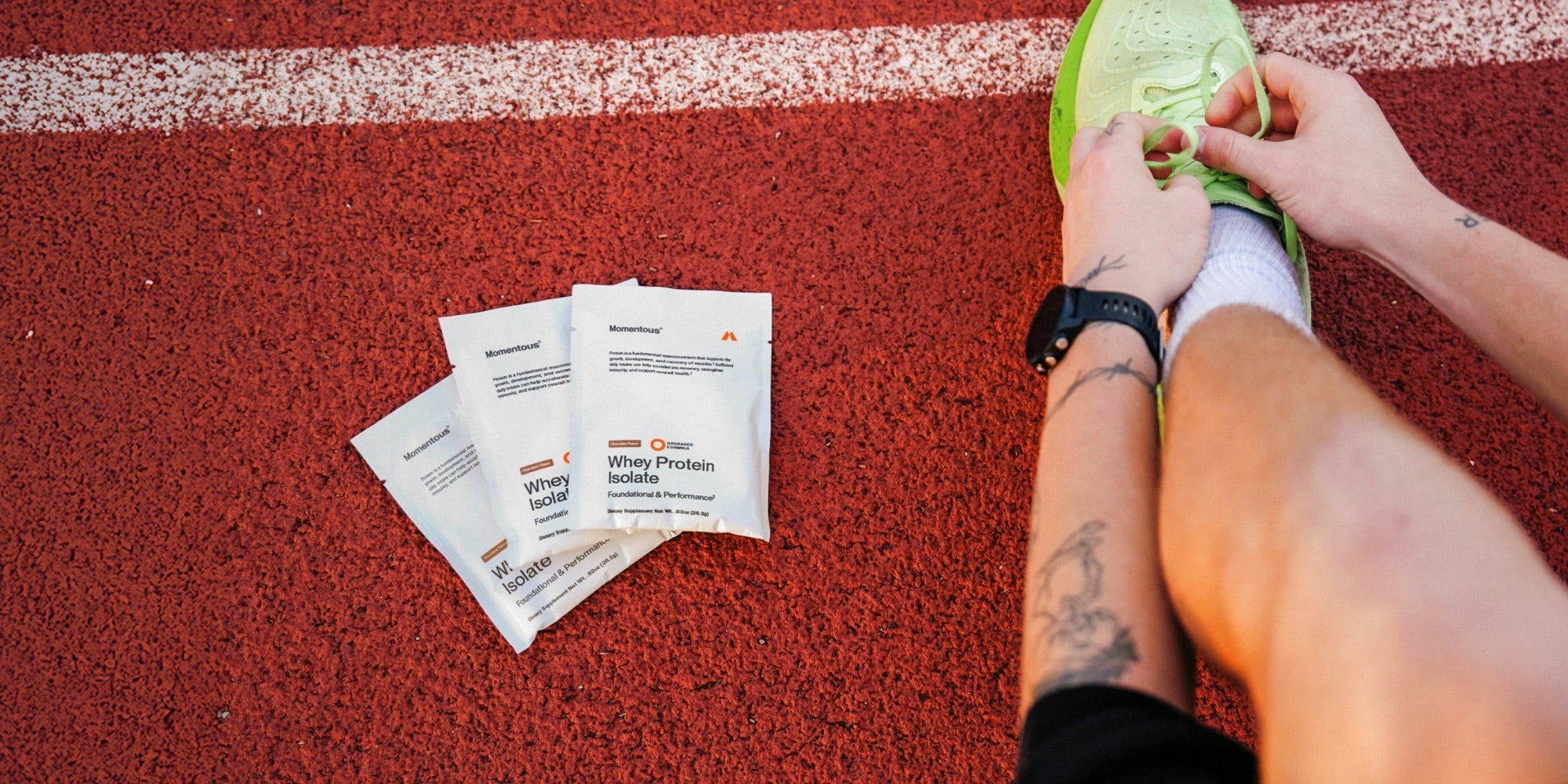Three travel packs of Momentous Whey Protein Isolate lay on the ground next to a runner's foot as he ties his shoelaces