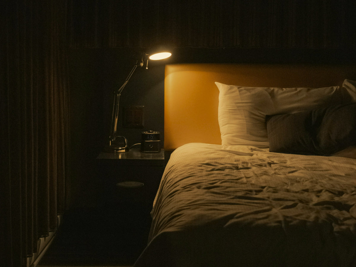 Dark bedroom at night, desk lamp lit and nightstand alarm clock reading 1:55 beside an unmade bed.