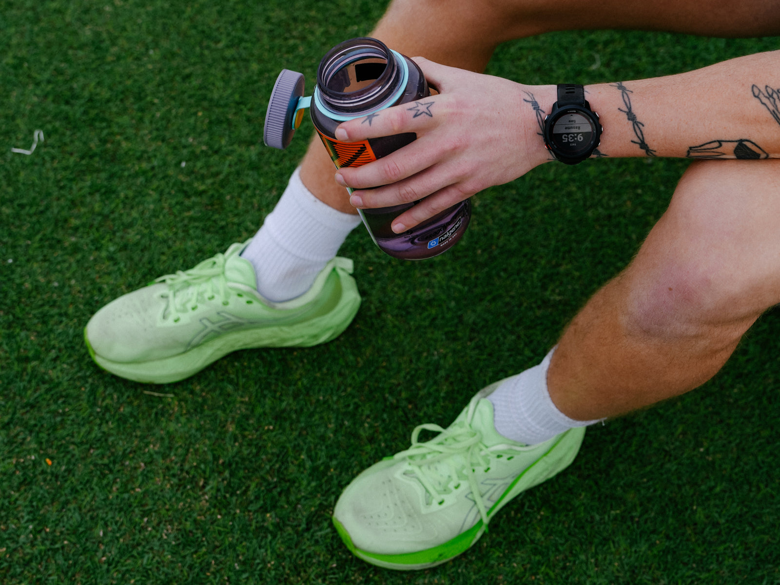 “Person sitting on grass holding a water bottle, wearing running shoes and watch.”
