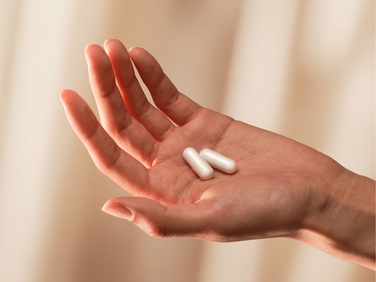 Open palm holding two white Momentous Magnesium Bisglycinate capsules against a soft beige curtain.