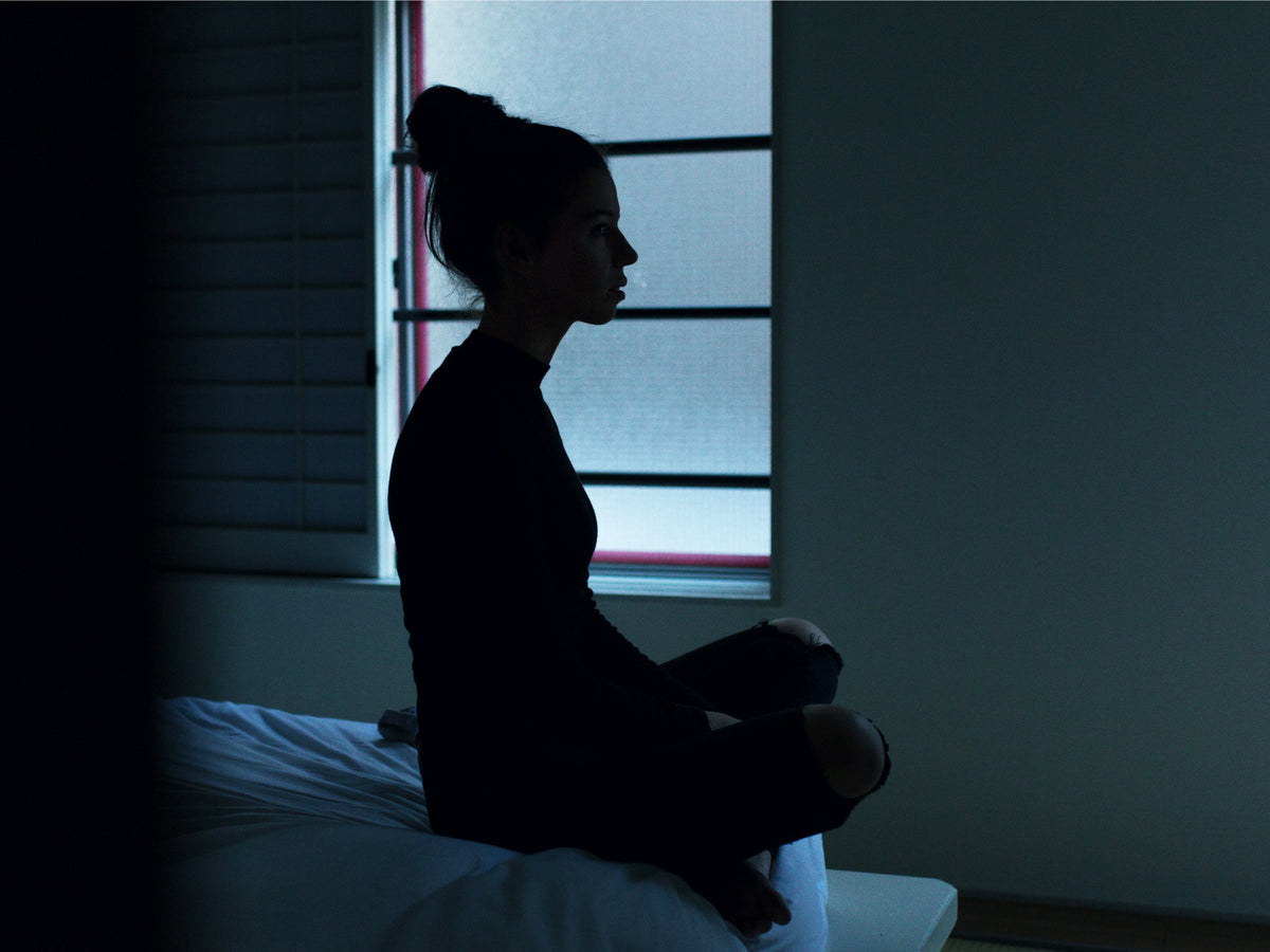 Person sitting cross-legged on the edge of a bed in a dark room, silhouetted against a window, awake at night.