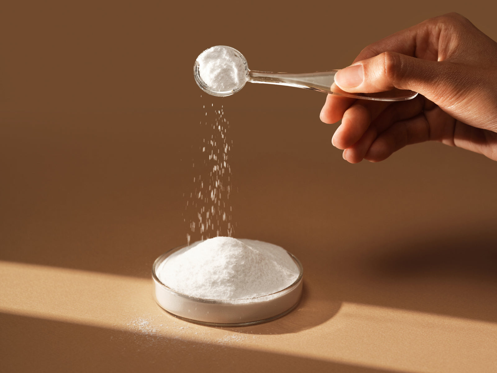 “Hand holding a scoop pouring white powder onto a small pile on a surface.”