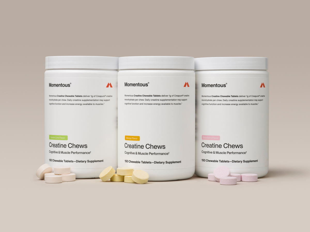 Three white containers of Momentous Creatine Chews are displayed side by side against a warm beige background, each with loose chews scattered in front. Left to right: Lemon Lime Flavor (green label), Mango Flavor (orange label), and Strawberry Flavor (pink label). Each label reads: "Creatine Chews – Cognitive & Muscle Performance, 150 Chewable Tablets – Dietary Supplement."