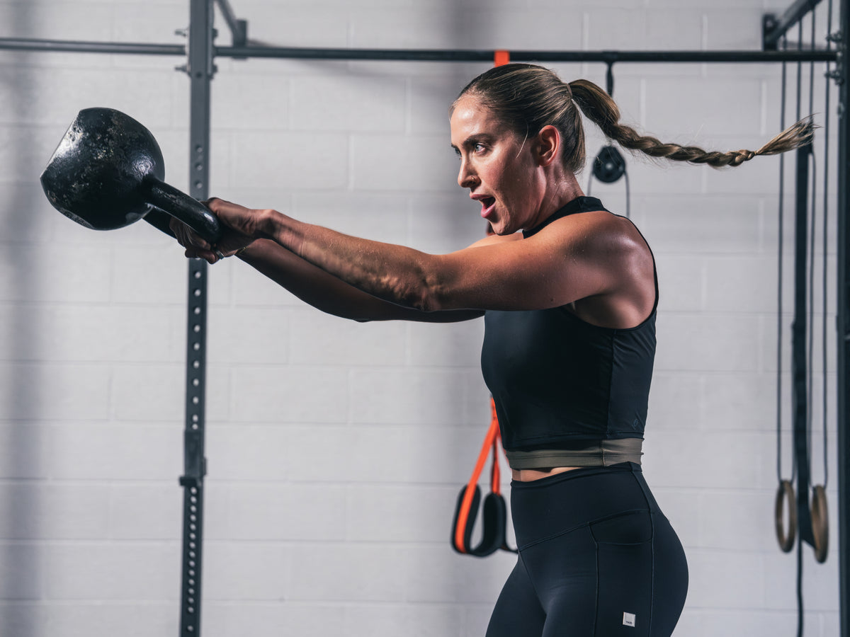 A female athlete in black workout gear performs a dynamic kettlebell swing in a gym setting. Her braided ponytail is in motion, conveying intensity and power. No text overlay.