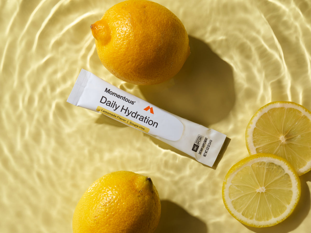 Momentous Daily Hydration stick pack, Lemonade Flavor, Single Use, NSF Certified Sport, Dietary Supplement, Net Wt 0.12oz 3.4g, lying among whole lemons and lemon slices in shallow water. (187 chars)