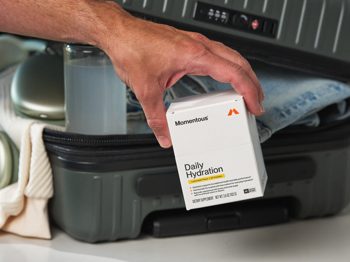 Momentous Daily Hydration box held above an open suitcase, Lemonade Flavor, 30 Packets: hydration supports foundational health and daily performance, intentionally formulated with less sodium for everyday hydration, naturally sweetened with honey and cane sugar with no artificial colors or flavors, NSF Certified Sport, Dietary Supplement, Net Wt 3.6oz 102g. (365 chars)