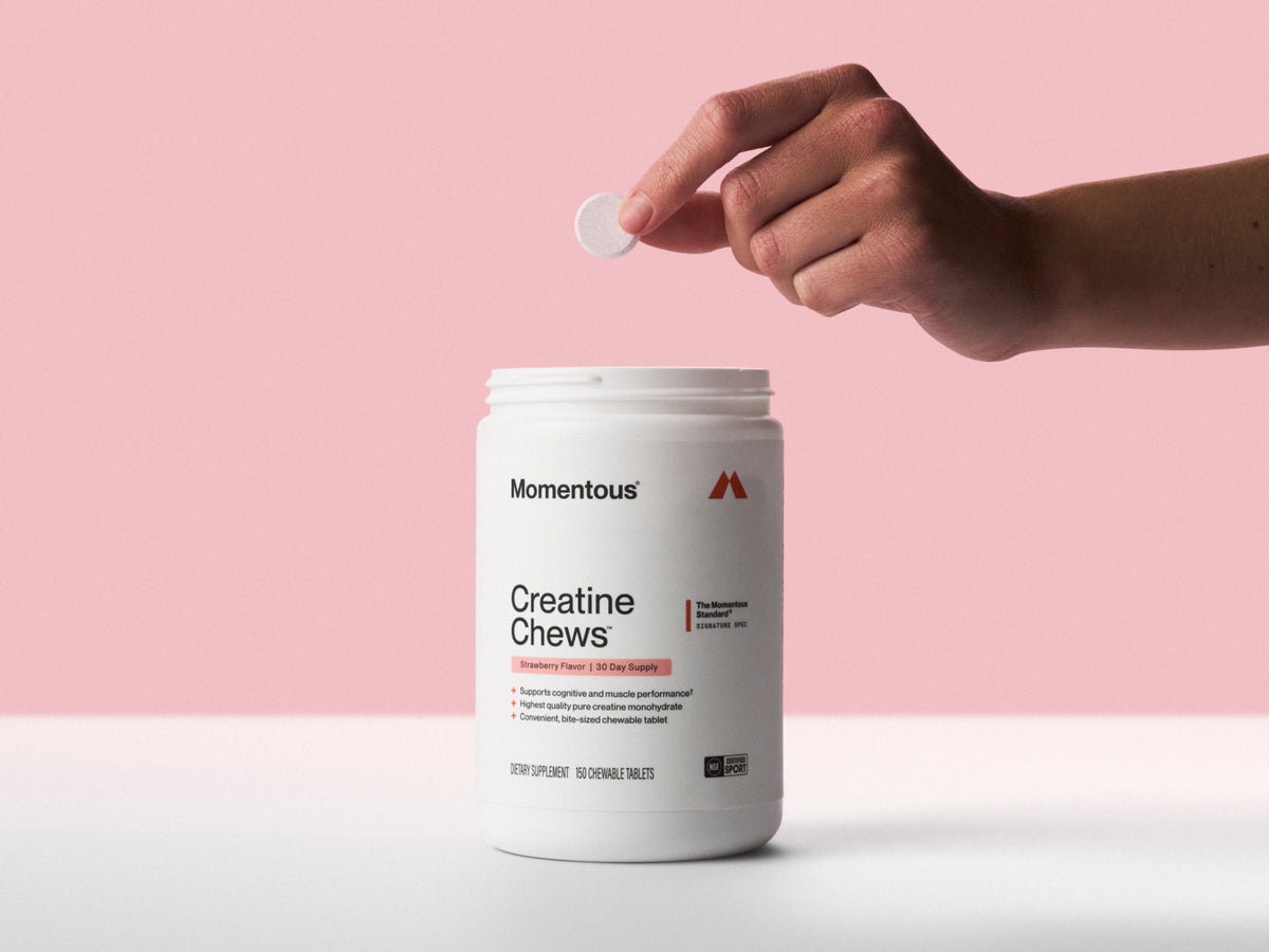 1. Hand holding a single chewable tablet above an open Momentous Creatine Chews tub on a pink backdrop, Strawberry Flavor, 30 day supply, 150 chewable tablets: supports cognitive and muscle performance, highest quality pure creatine monohydrate, convenient bite-sized chewable tablet, The Momentous Standard Signature Spec, dietary supplement, NSF Certified Sport.  (361 chars)