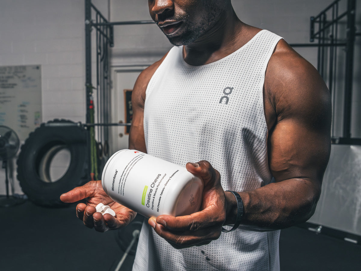 A muscular athlete in a white mesh tank top holds a container of Momentous Creatine Chews, tilting it to pour chewable tablets into his open palm. The label reads: "Creatine Chews – Cognitive & Muscle Performance, 90 Chewable Tablets – Dietary Supplement." Set in a gym environment.