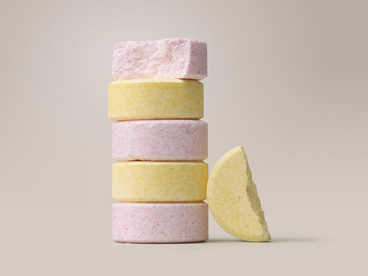 A styled close-up of five chewable tablets stacked vertically, alternating between pink and yellow, against a warm beige background. A broken yellow tablet leans against the stack. No text overlay.