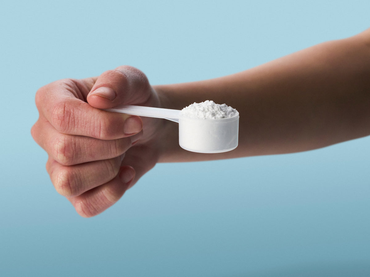 A hand holds a white measuring scoop filled with fine white powder against a soft blue background.