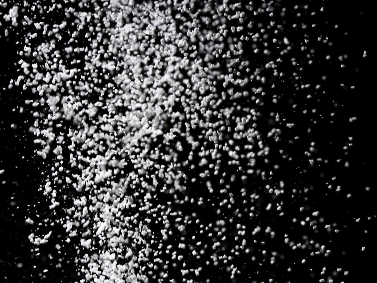 A dramatic close-up of fine white powder particles suspended mid-air against a solid black background, captured in high contrast black and white.
