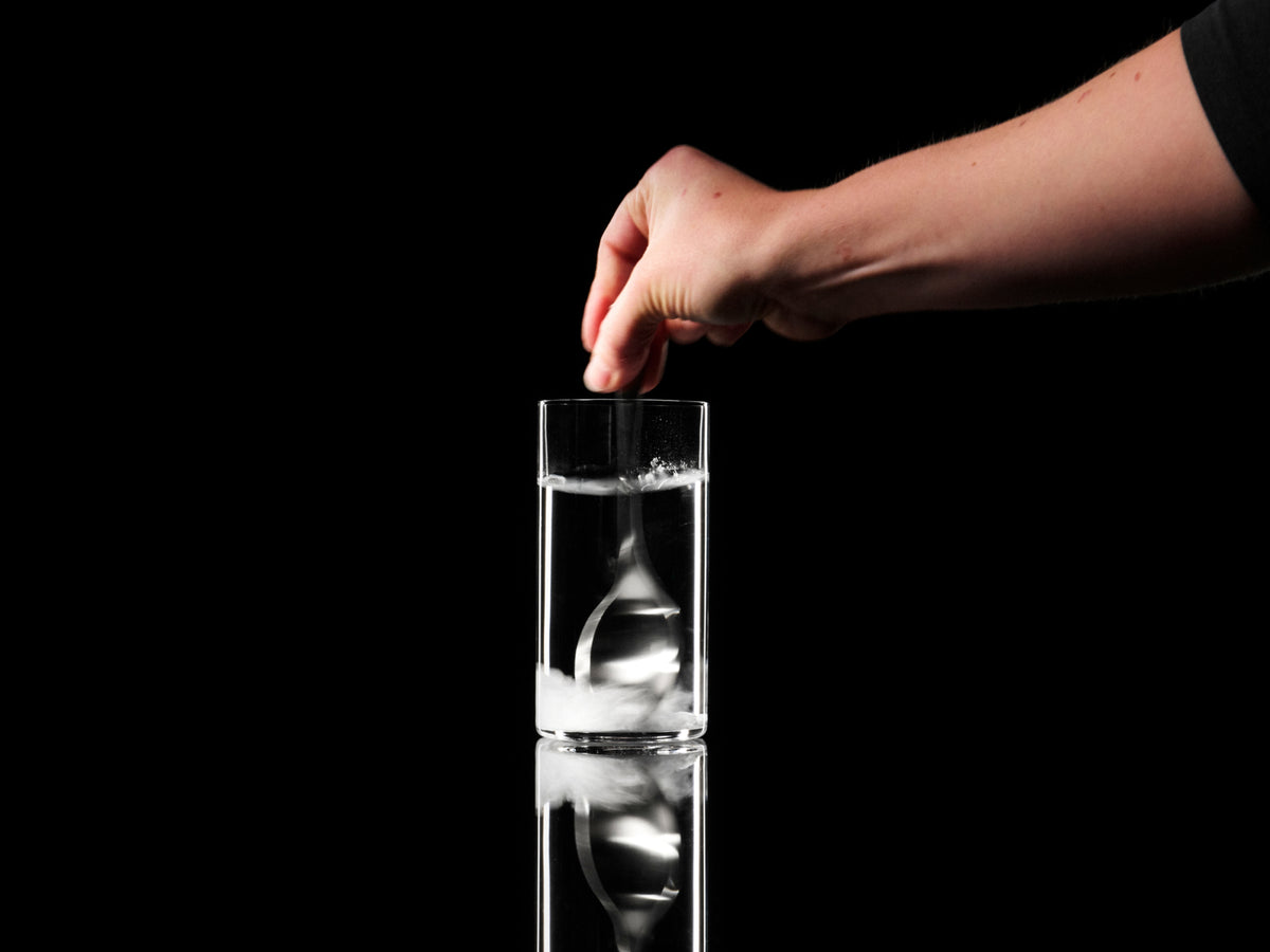 A hand drops a pinch of white powder into a tall clear glass of water against a black background, creating a swirling white cloud as it dissolves.