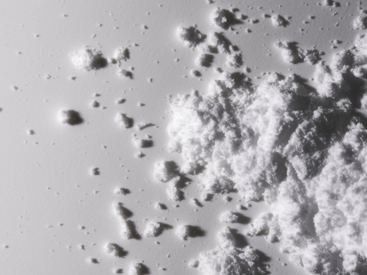 An extreme close-up of fine white powder granules scattered and piled on a light gray surface, with detailed texture and soft shadows visible.
