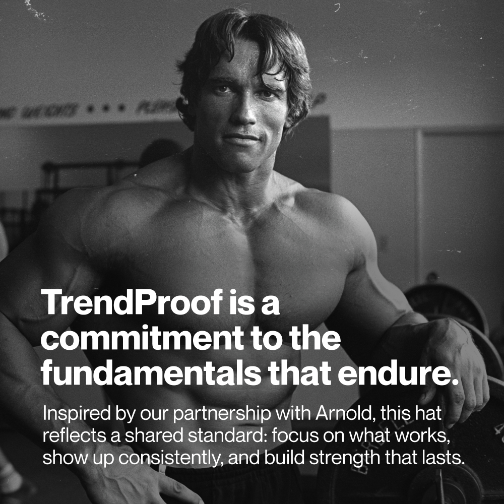 Black-and-white photo of Arnold Schwarzenegger in a gym environment. Large white overlay text reads: “TrendProof is a commitment to the fundamentals that endure.” Smaller text below reads: “Inspired by our partnership with Arnold, this hat reflects a shared standard: focus on what works, show up consistently, and build strength that lasts.” Arnold is shown shirtless holding a weight plate with gym equipment visible in the background.