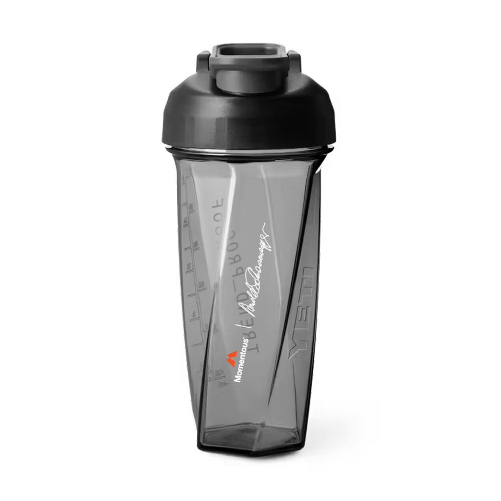 Front view of a transparent gray Helimix shaker bottle with a black screw-top lid and flip cap. The bottle has a faceted, geometric shape with embossed measurement markings on the side. Printed vertically on the front is a white Arnold Schwarzenegger signature above a small orange Momentous logo and white “Momentous” wordmark. The bottle appears designed for mixing supplements or protein shakes.