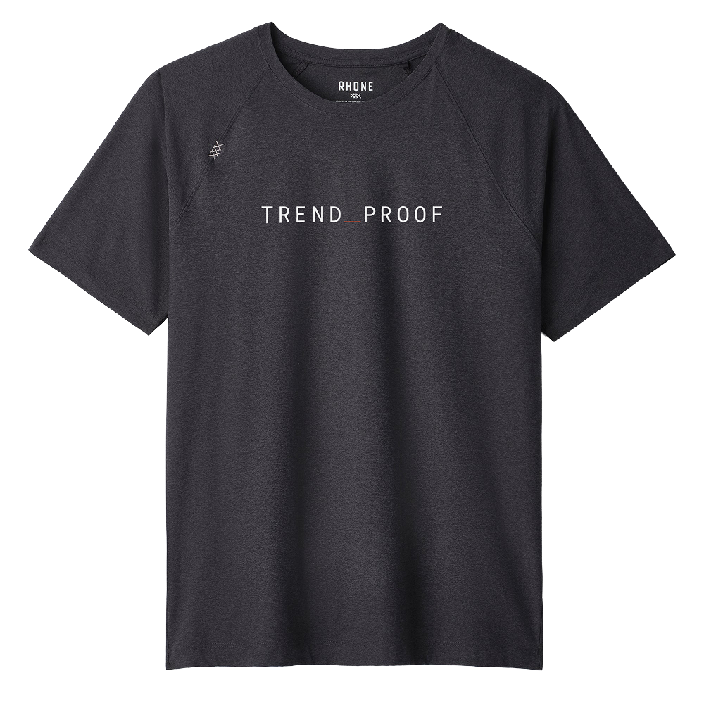 Front view of a charcoal gray short-sleeve performance T-shirt with raglan sleeves. The shirt features a minimalist design with the word “TREND_PROOF” printed in clean white uppercase lettering across the chest, with a subtle red underscore between the words. A small stitched emblem detail appears near the left shoulder seam. The collar label reads “RHONE.” The fabric appears lightweight and athletic with a smooth, structured drape.