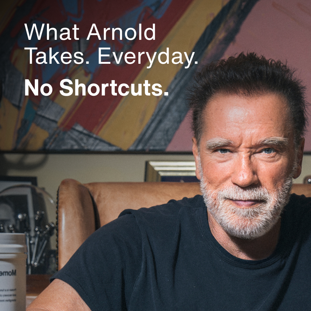 Arnold Schwarzenegger seated indoors with a Momentous product visible beside him. Text reads: What Arnold Takes. Everyday. No Shortcuts.