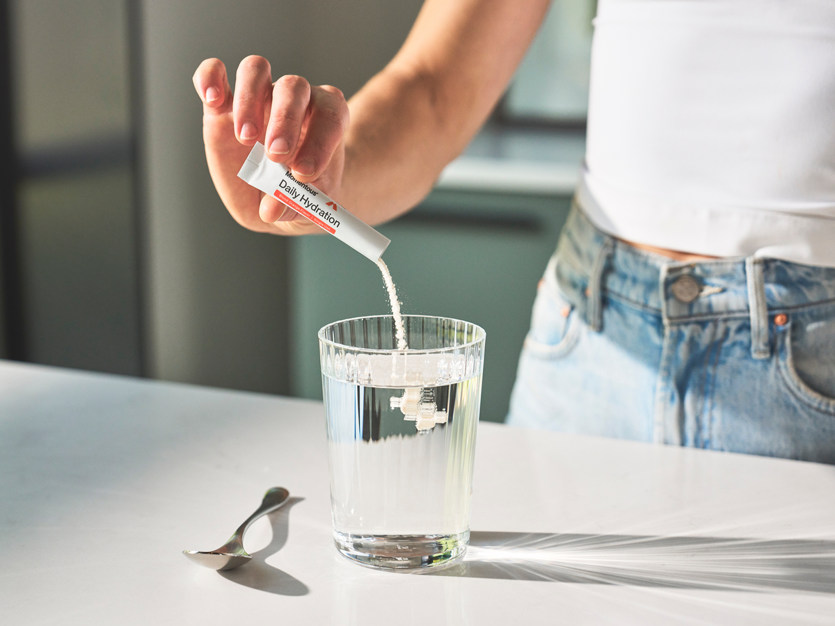 Hand pouring a Momentous Daily Hydration stick pack, Blood Orange Flavor, Single Use, into a glass of water on a white counter, with a metal spoon beside it. (161 chars)