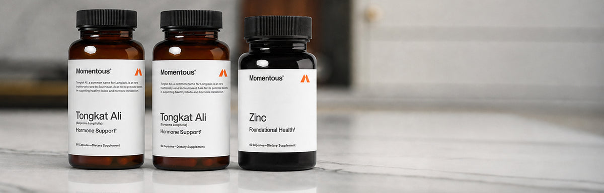 Three supplement bottles on a marble surface with a blurred neutral interior background. Left to right: two amber glass bottles labeled Momentous Tongkat Ali (Eurycoma Longifolia), "Hormone Support," 60 Capsules—Dietary Supplement; and one dark bottle labeled Momentous Zinc, "Foundational Health," 60 Capsules—Dietary Supplement. Each label has the orange Momentous "M" logo in the top right.