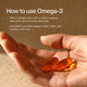 A hand holds two amber omega-3 softgel capsules against a warm-toned background. Text on the image reads: “How to use Omega-3” “• Take 2 softgels daily with a meal to support absorption and minimize aftertaste.” “• Consistent use helps support heart, brain, and recovery over time.”